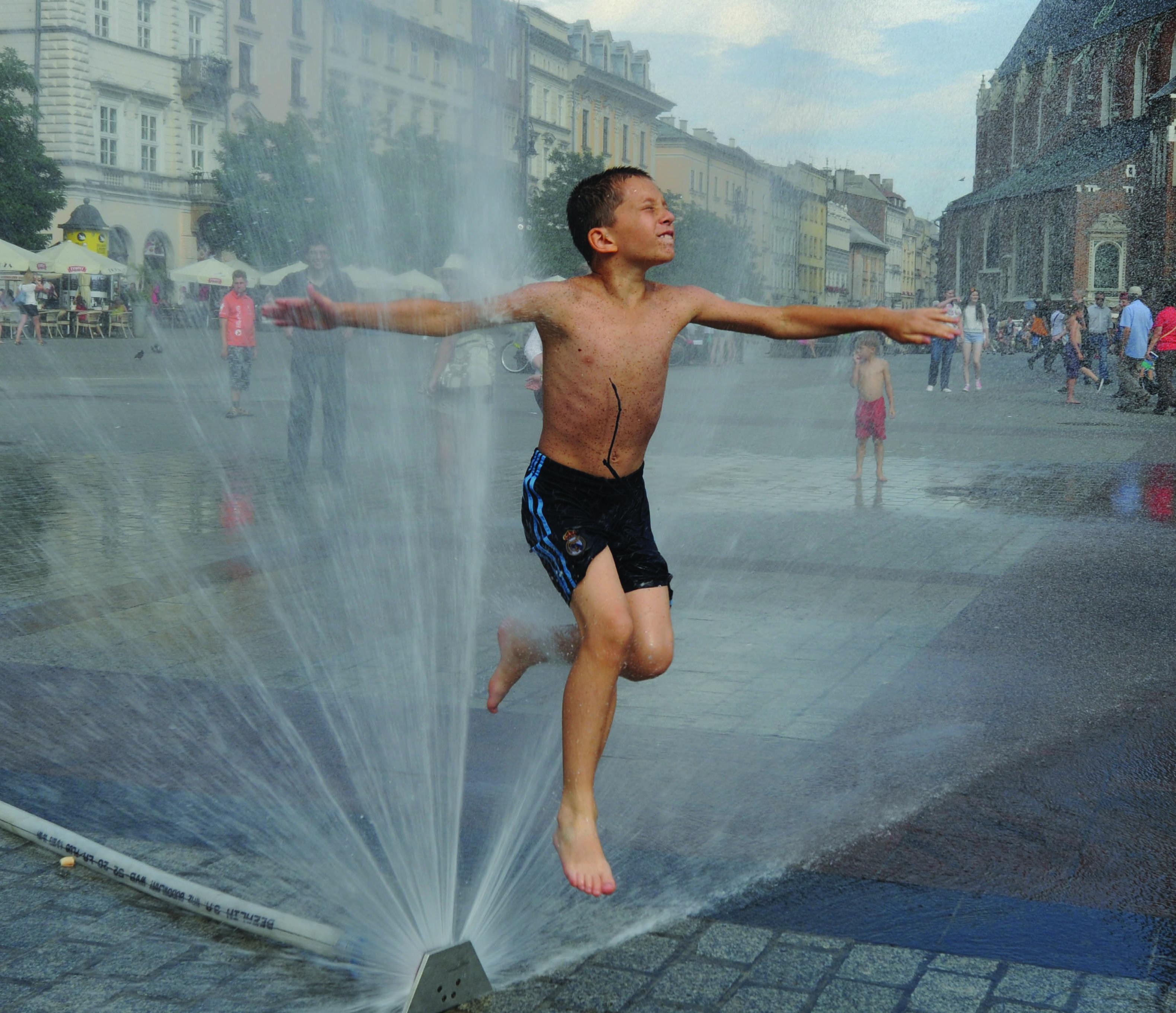 Fotografia na rua de criança em calções de banho a saltar por cima de repuxo}
