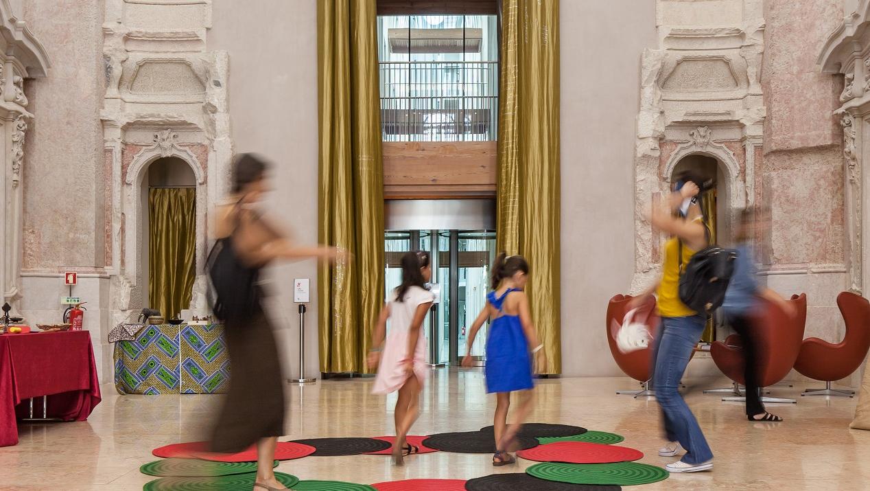 Duas mulheres e duas crianças andam à volta sob tapetes de várias cores (vermelho, verde e preto)